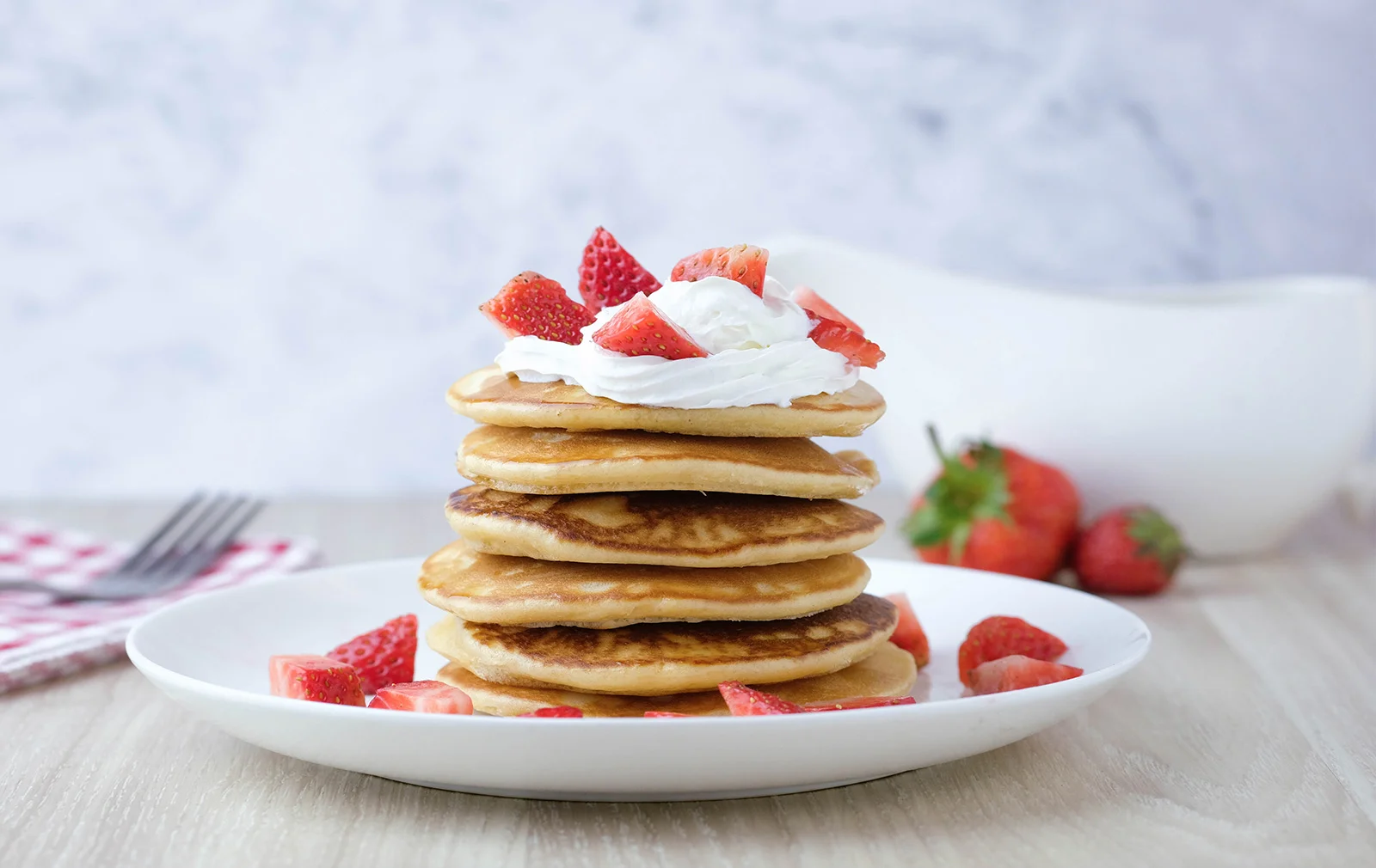 Pancakes caseros con crema vegetal para batir y fresas frescas, servidos en un plato blanco sobre una mesa.